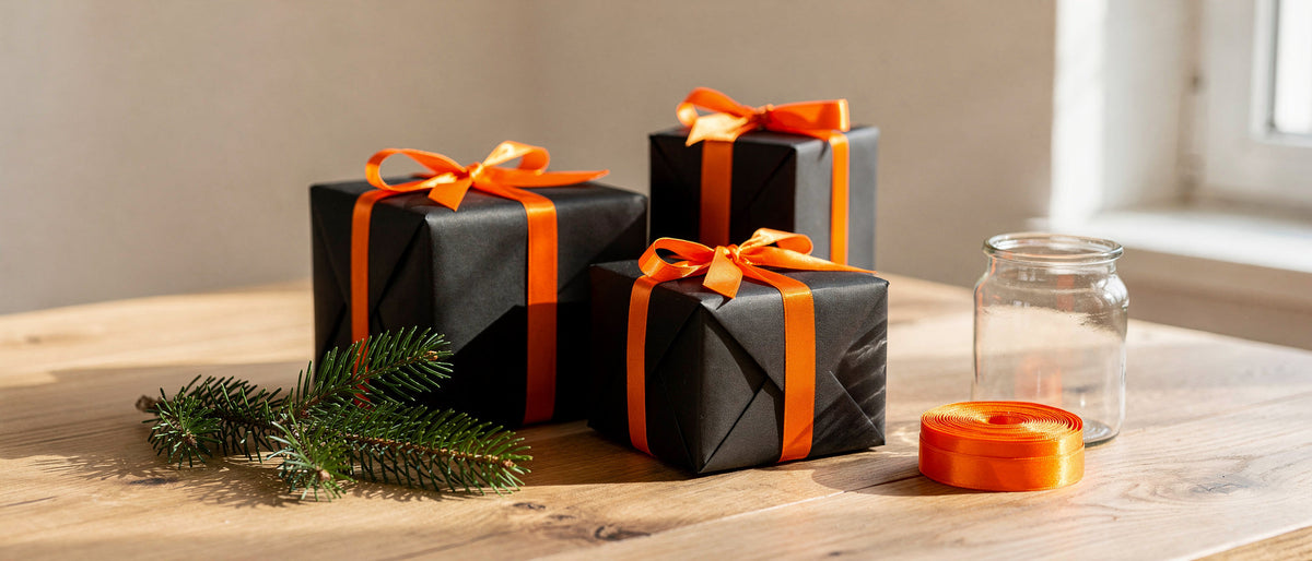 Drei schwarz verpackte Geschenke mit orangefarbenen Schleifen auf einem Holztisch, daneben Tannenzweig und Glas